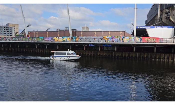 A small white and blue passenger ferry boat traveling along a urban river with a boardwalk wall covered in colorful street art graffiti and large stad
